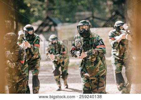 Selective Focus Of Paintball Team In Uniform And Protective Masks Playing Paintball With Marker Guns