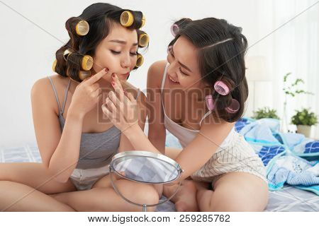 Two Young Asian Women Looking At Mirror And Poking At Cheek While Sitting In Bedroom Together