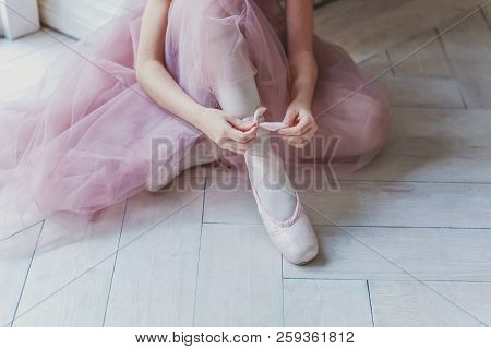 Hands Of Ballerina In Pink Tutu Skirt Puts On Pointe Shoes On Leg In White Light Hall. Young Classic