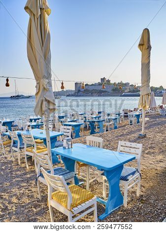 Tables With Chairs Of A Turkish Tavern In A Beach Of Kumbahce Bay With The Castle Of Saint Peter In