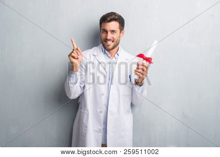 Handsome young doctor man over grey grunge wall holding diploma very happy pointing with hand and finger to the side