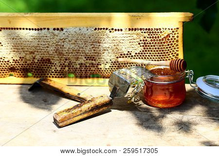 Jar Of Fresh Honey In A Glass Jar, Beekeeping Tools Outside. Frame With Bees Wax Structure Full Of F