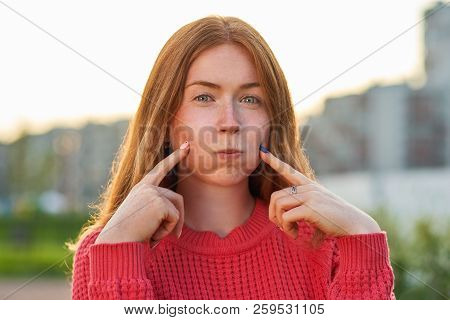 A Funny Red-haired Girl Inflates Her Cheeks And Pokes Her Index Fingers At Them. The Concept Of Fun,