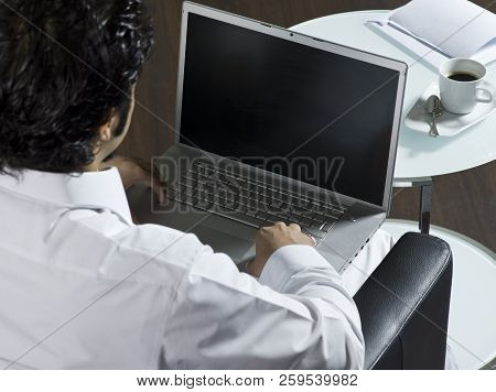 Mideastern Man Using Laptop At Office, With A Cup Of Cofee And Documents On The Side Table