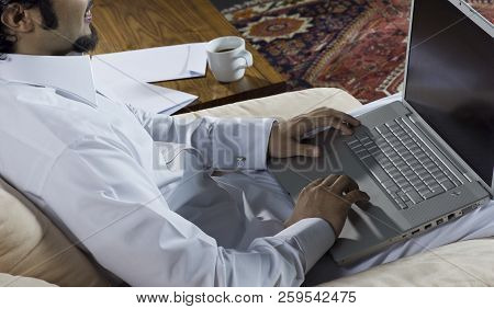 Mideastern Man Using Laptop At Office, With A Cup Of Cofee And Documents On The Side Table