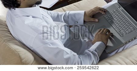 Mideastern Man Using Laptop At Office, With A Cup Of Cofee And Documents On The Side Table