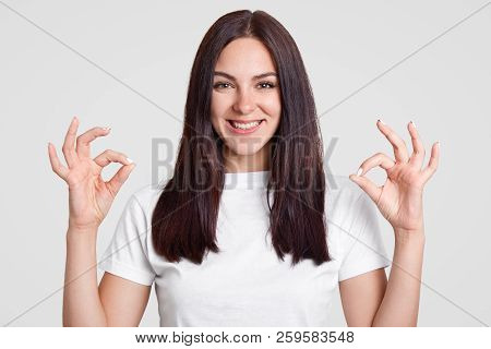 Indoor Shot Of Happy Attractive Woman With Long Straight Dark Hair, Makes Okay Sign With Both Hands,