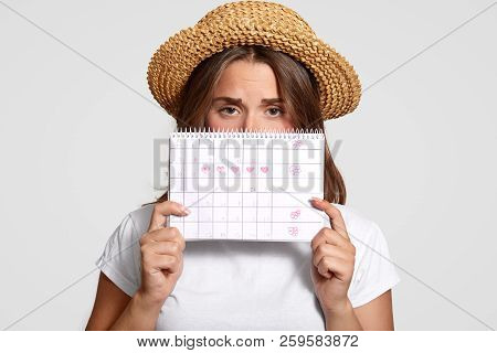 Displeased Woman Holds Perids Calendar, Wears Straw Hat, Has Discontent Expression, Dressed In Casua