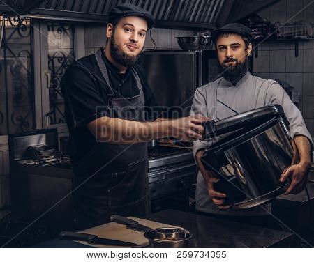 Two Brutal Cooks Dressed In Uniforms And Hats Standing With Big Multicooker In The Kitchen.