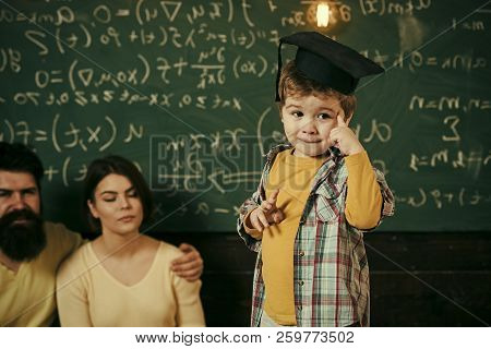 Smart Child, Wunderkind In Graduate Cap Pointing At His Head. Boy Presenting His Knowledge To Mom An