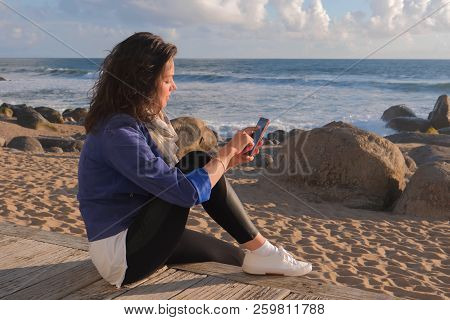 Beautiful Black Hair Woman Sitting On Wooden Embankment And Using Smartphone