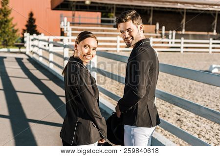 Back View Of Smiling Equestrians In Professional Apparel Standing At Ranch And Looking At Camera