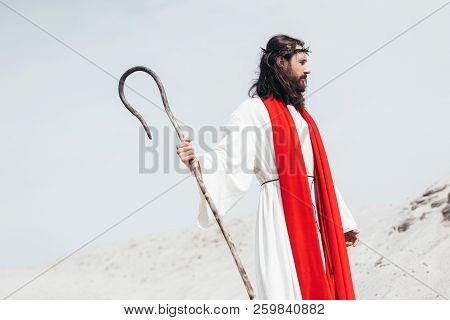 Side View Of Jesus In Robe, Red Sash And Crown Of Thorns Standing With Wooden Staff In Desert