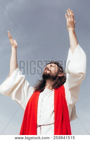 Low Angle View Of Jesus In Robe, Red Sash And Crown Of Thorns Standing With Raised Hands Against Gre