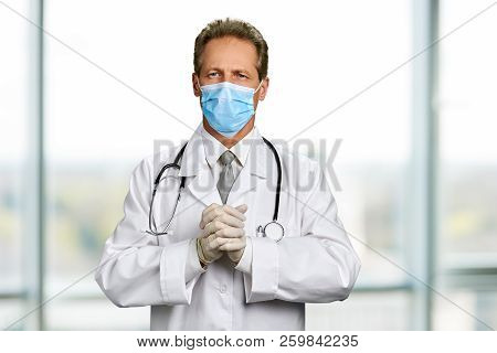Doctor In Gloves Clasps His Hands Together. Mature Medical Doctor In White Coat And Face Mask Standi