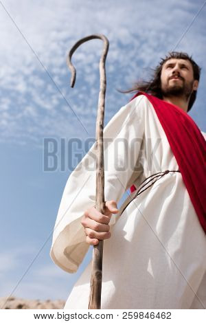 High Angle View Of Jesus In Robe, Red Sash And Crown Of Thorns Standing In Desert With Wooden Staff