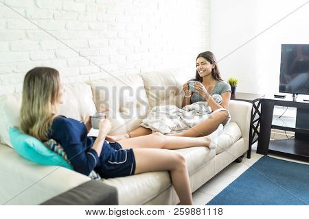 Happy Young Woman Holding Coffee Cup While Chitchatting With Friend On Sofa At Home