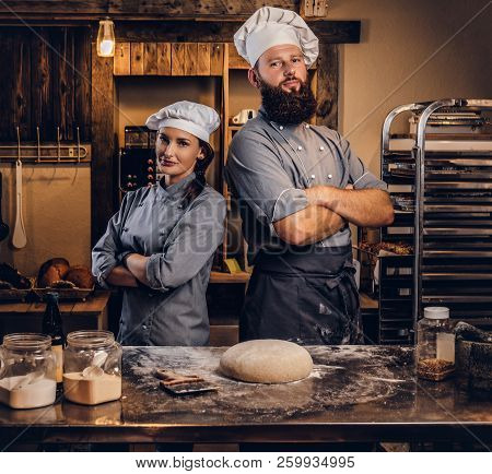 Chef With His Assistant In Cook Uniform Posing With Crossed Arms Near Table With Ready Dough In The 