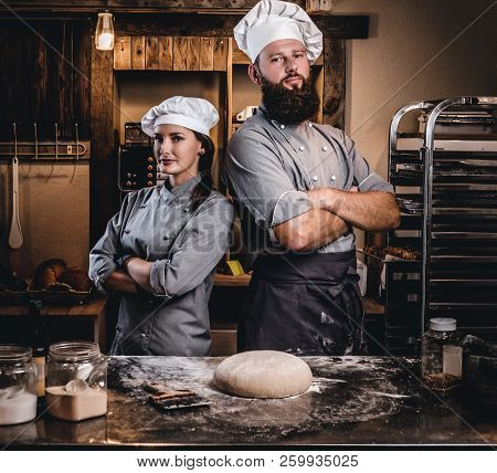 Chef With His Assistant In Cook Uniform Posing With Crossed Arms Near Table With Ready Dough In The 