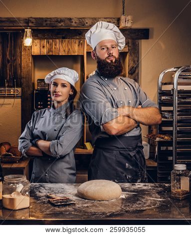 Chef With His Assistant In Cook Uniform Posing With Crossed Arms Near Table With Ready Dough In The 