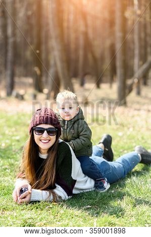 Happy Mom And Toddler Son Spend Time Outdoors. The Kid With Blond Hair Sits On The Back Of His Mothe