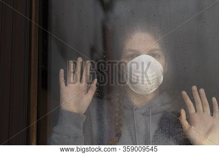 Young Girl Wearing A Mask Is Looking With Hope The Window On Which Raindrops, Is Staying At Home Iso