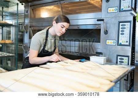 Focused Female Baker Cutting Open Some Loaves Of Bread Before Baking Them In The Oven