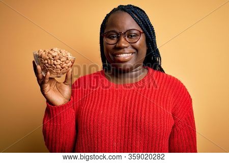 Young african american plus size woman with braids holding bowl with healthy peanuts with a happy face standing and smiling with a confident smile showing teeth