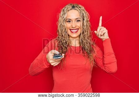 Young beautiful blonde woman using television remote control over isolated red background surprised with an idea or question pointing finger with happy face, number one