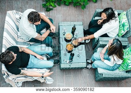 High Angle Of Four Friends Sitting Together On Terrace, Drinking Wine, Eating Snacks And Chatting. Y