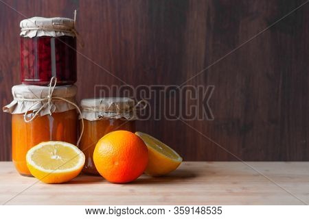 Jars With Various Jams And Lemon Cut And Orange On Dark Wooden Background. Strowberry And Apricot Ja
