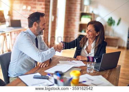 Two middle age business workers smiling happy and confident. Working together with smile on face using laptop doing handsake at the office