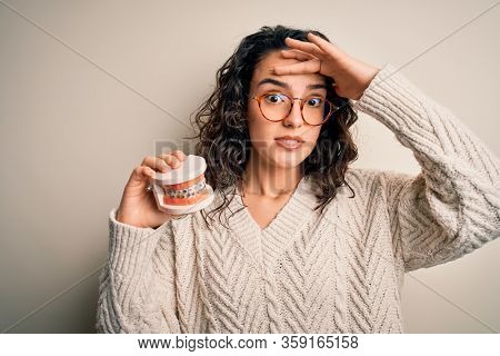 Young beautiful woman with curly hair holding plastic teeth with dental baces stressed with hand on head, shocked with shame and surprise face, angry and frustrated. Fear and upset for mistake.