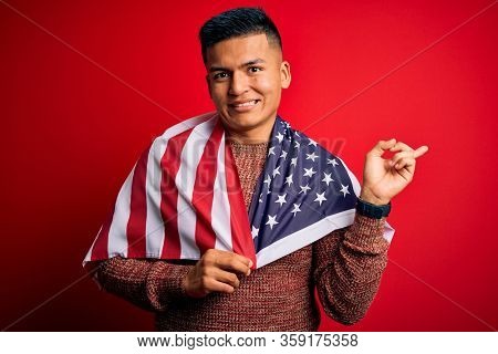 Young handsome latin patriotic man wearing united states of america falg very happy pointing with hand and finger to the side