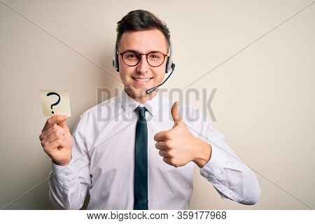 Young call center operator man wearing headset holding paper note with question mark happy with big smile doing ok sign, thumb up with fingers, excellent sign