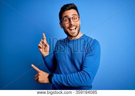 Young handsome man with beard wearing casual sweater and glasses over blue background smiling and looking at the camera pointing with two hands and fingers to the side.