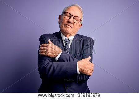 Grey haired senior business man wearing glasses and elegant suit and tie over purple background Hugging oneself happy and positive, smiling confident. Self love and self care