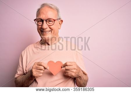 Senior grey haired man holding heart shape paper over pink background with a happy face standing and smiling with a confident smile showing teeth