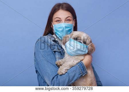 Horizontal Shot Of Woman Wearing Denim Jacket, Face Mask And Hoding Dog With Flumask Too, Posing Iso