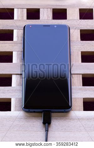 Brecht, Belgium - April 1 2020: A Smartphone Lying On A Wooden Box With A Usb Cable In Its Connector