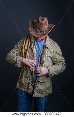 Cowboy In A Wide-brimmed Hat And Jacket With A Lasso On His Shoulder Posing On A Dark Background