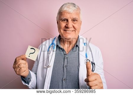 Senior handsome hoary doctor man wearing stethoscope holding reminder with question mark happy with big smile doing ok sign, thumb up with fingers, excellent sign