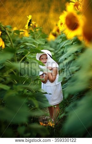 A Cute Little Smiling Girl In The Field Of Sunflowers. Child In Momy Hat. Childhood Concept. Sunny S