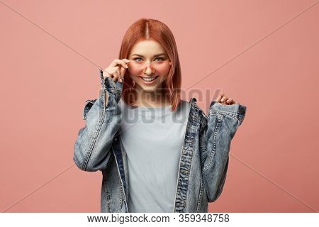Smiling ginger woman in pink glasses in denim jacket standing in studio