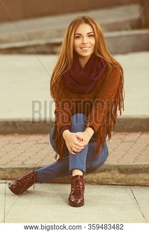 Happy young fashion woman sitting on street sidewalk  Stylish female model in brown leather fringe suede jacket and dark blue jeans