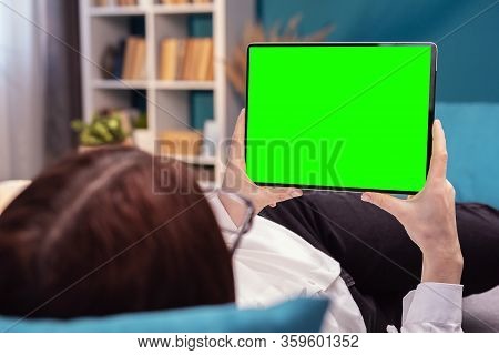 Back View Of Female Holding Horizontal Tablet With Green Screen Lying On Bed Or Sofa In Apartment
