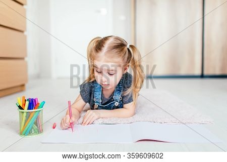 Blonde Happy Little Girl With Two Ponytales Drawing And Writing Lying On The Floor At Home. Preschoo