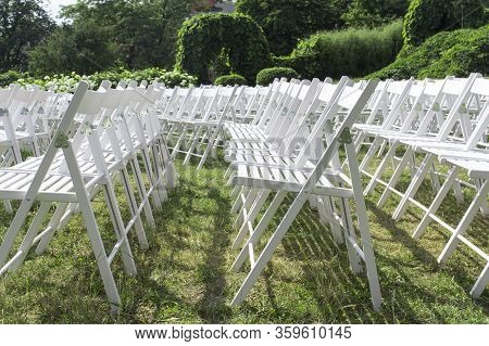 View Of Empty Wooden Chairs In White, Arranged In Rows, Against The Background Of Hedges And Climbin