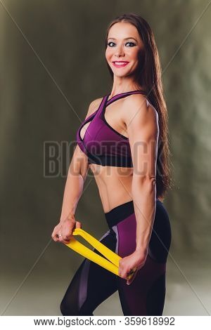 Athletic Girl Performs Exercises Using A Resistance Band. Photo Of Young Girl On Drey Background. St