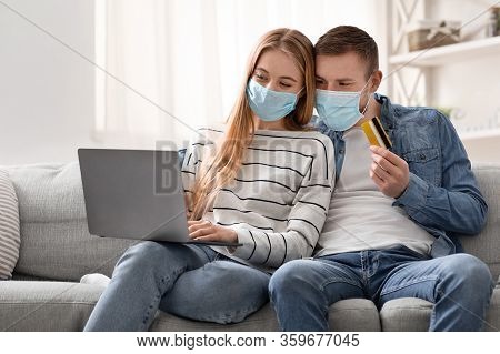 Shopping Online During Quarantine. Young Couple In Protective Masks Purchasing In Internet With Cred